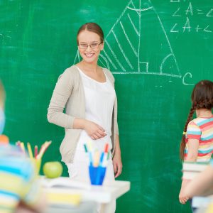 Portrait of smart teacher standing by blackboard and looking at schoolkids while one of pupils standing near by